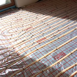 UBS Usługi Budowlane Stępień Lech Stępień - Równolegle ułożone rury ogrzewania podłogowego marki Roth na folii z czerwoną siatką, widoczne mocowania rur i fragment okna.