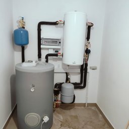 UBS Usługi Budowlane Stępień Lech Stępień - Zestaw zbiorników buforowych i instalacji w kotłowni, widoczne czarne rury izolowane, białe ściany i brudna podłoga z płytek.