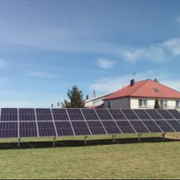 Rząd paneli fotowoltaicznych na metalowym stelażu, zainstalowany na trawiastym terenie przed domem z czerwoną dachówką, błękitne niebo z lekkimi chmurami w tle.