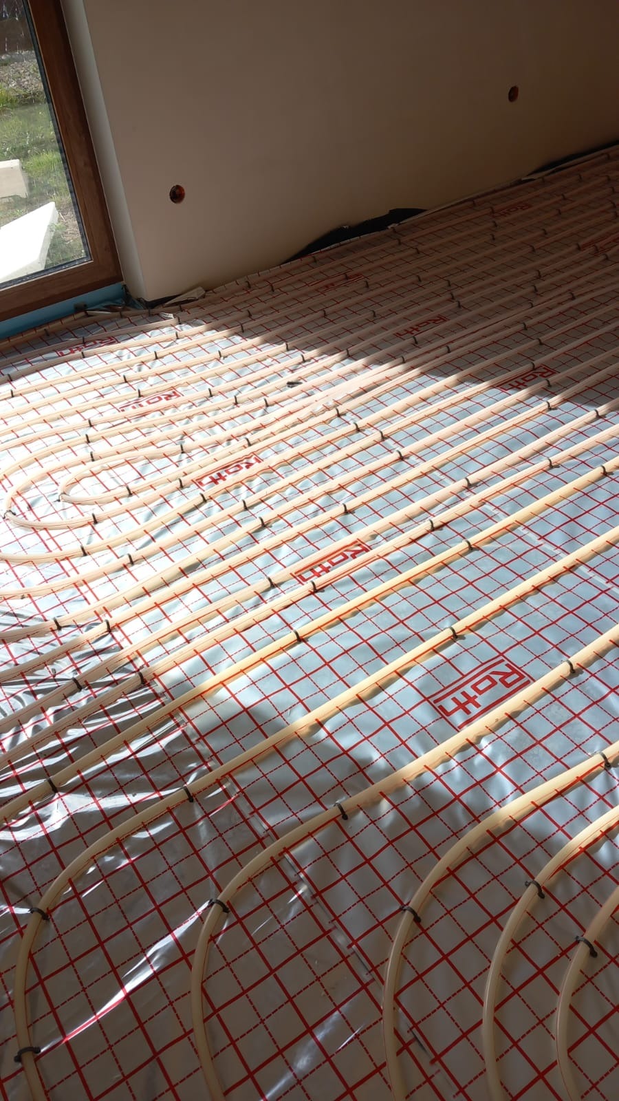 Równolegle ułożone rury ogrzewania podłogowego marki Roth na folii z czerwoną siatką, widoczne mocowania rur i fragment okna.