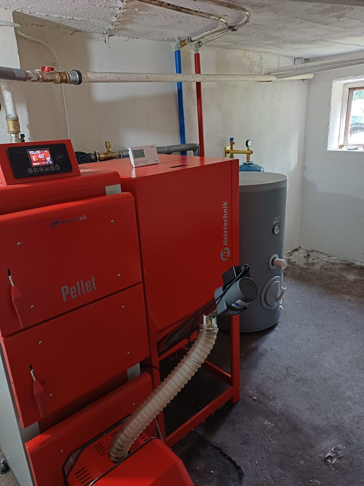 Czerwony piec na pellet marki Heiztechnik w kotłowni, z widocznym zbiornikiem akumulacyjnym i orurowaniem na ścianie.