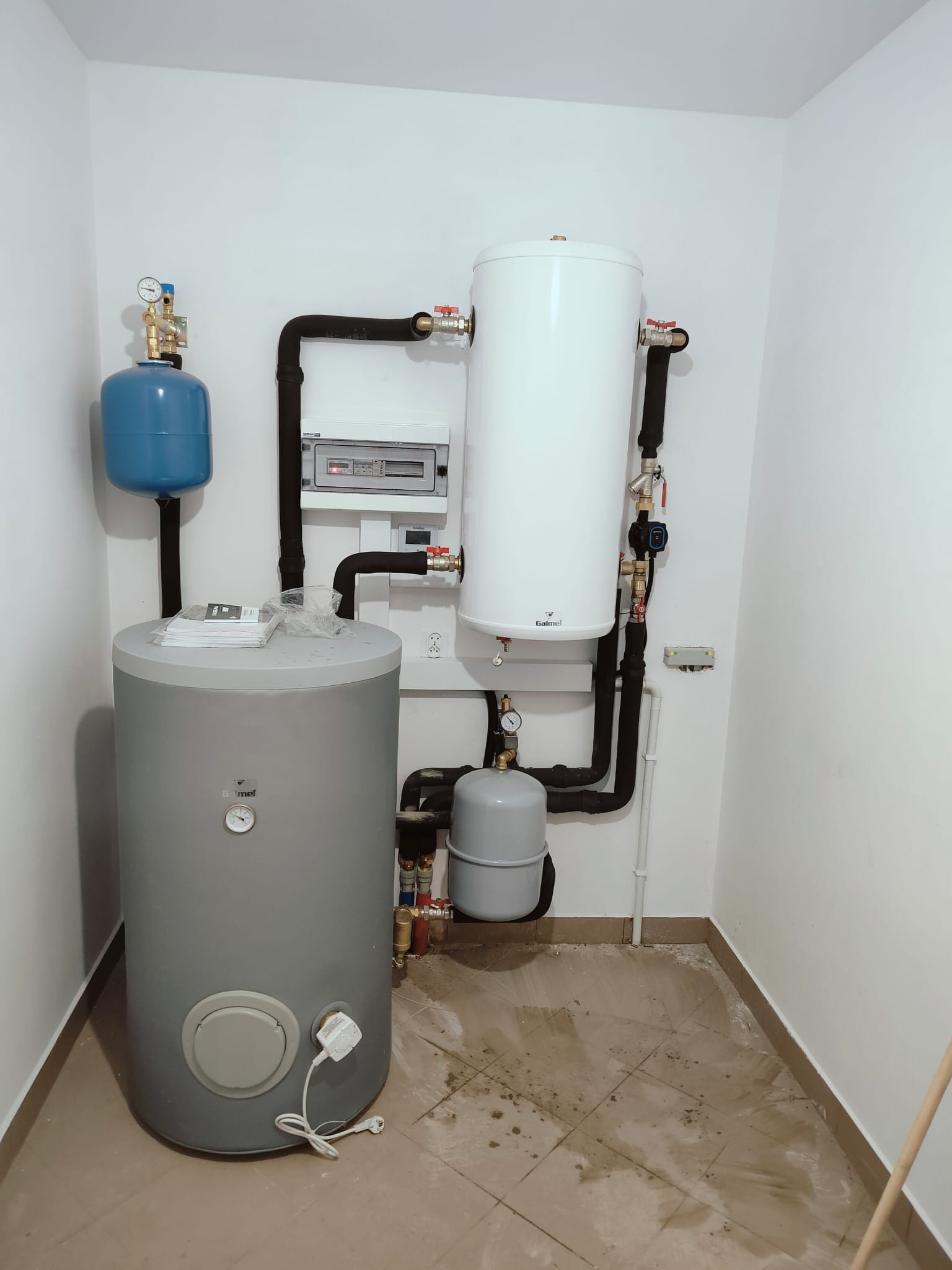 Zestaw zbiorników buforowych i instalacji w kotłowni, widoczne czarne rury izolowane, białe ściany i brudna podłoga z płytek.