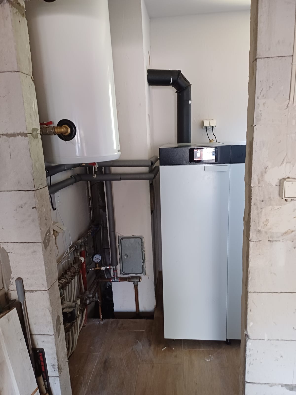 Nowoczesny piec gazowy z zasobnikiem ciepłej wody, zainstalowany w pomieszczeniu w Przasnyszu, widoczne rury i przyłącza, fragment ściany z niedokończonym wykończeniem.