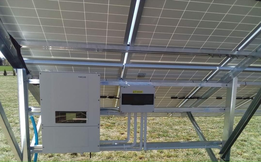 Widok od spodu na zamontowane panele fotowoltaiczne na metalowej konstrukcji, widoczny inwerter Sofar i skrzynka elektryczna z przewodami, na tle zielona trawa i drzewa.