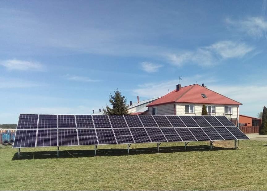 Rząd paneli fotowoltaicznych na metalowym stelażu, zainstalowany na trawiastym terenie przed domem z czerwoną dachówką, błękitne niebo z lekkimi chmurami w tle.