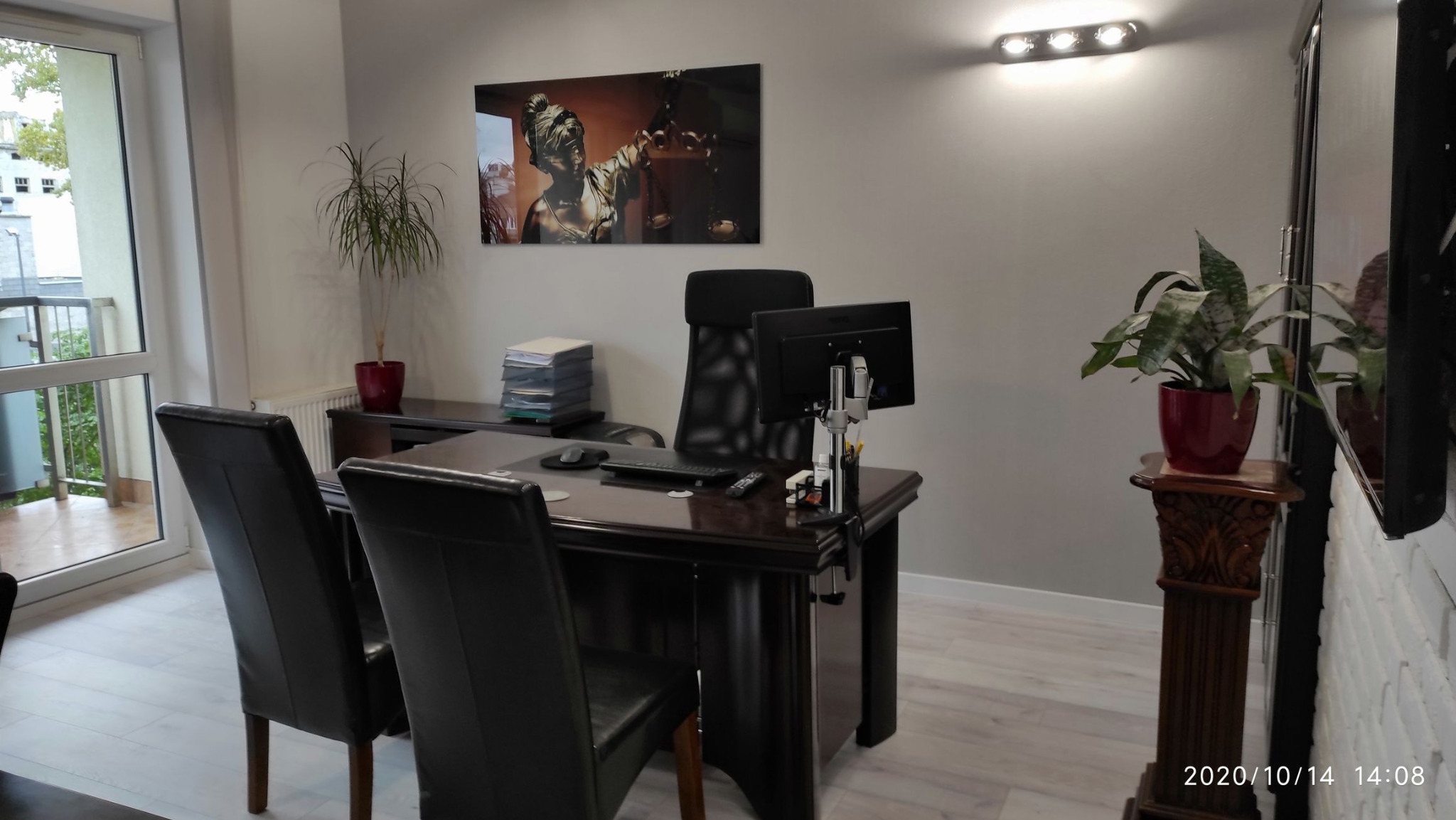 Biuro z ciemnym drewnianym biurkiem, czarnymi krzesłami i obrazem Temidy na ścianie, roślina doniczkowa na postumencie i dokumenty w tle.