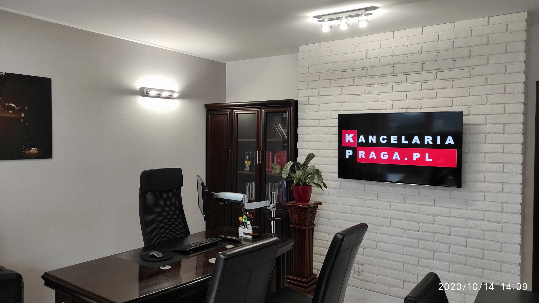 Wnętrze biura: ciemne meble, biała ściana z cegły, telewizor z napisem Kancelaria Praga.pl, roślina doniczkowa na kolumnie.