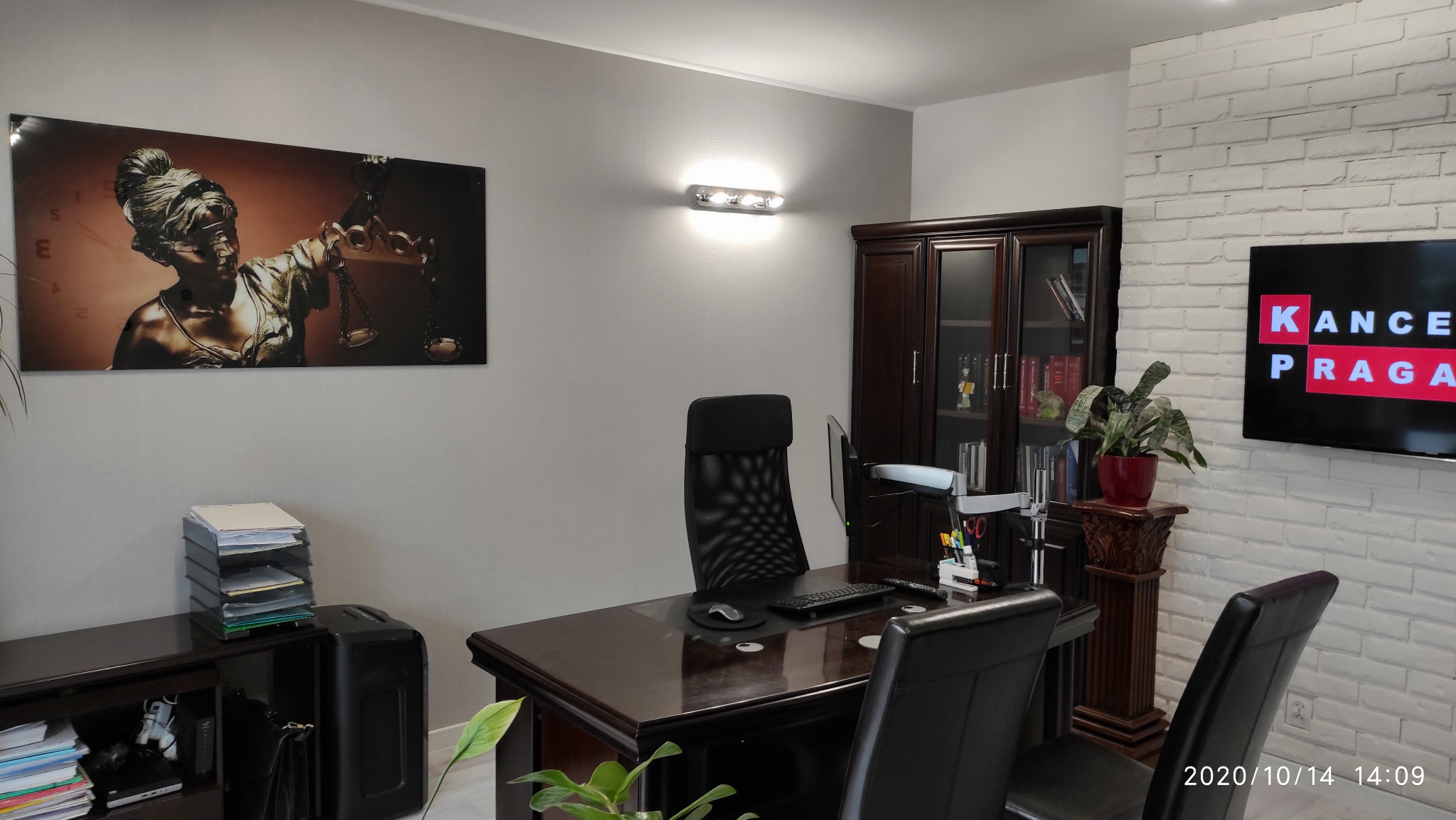 Wnętrze gabinetu z biurkiem, krzesłami i regałem. Na ścianie obraz z Temidą i telewizor z napisem 'KANCE PRAGA'.