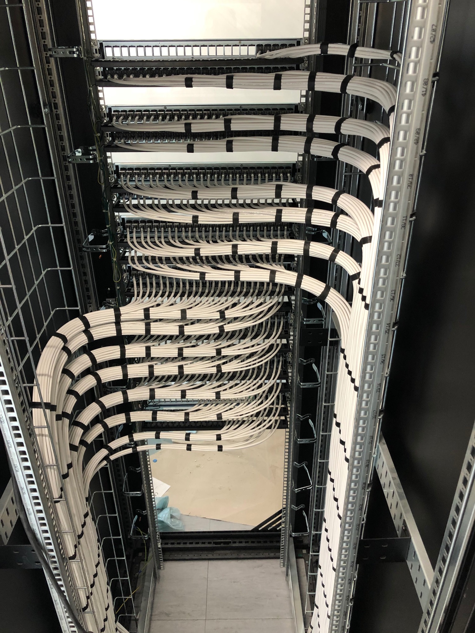 Rząd białych kabli uporządkowanych w metalowej szafie rack, zabezpieczonych czarnymi opaskami zaciskowymi i ułożonych w regularne łuki.