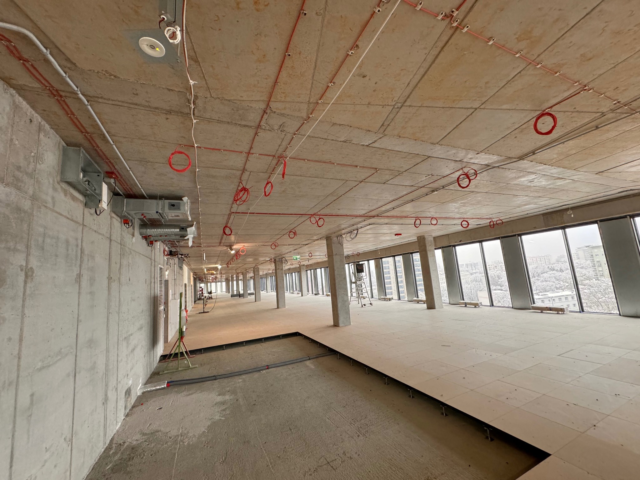 Wnętrze biurowe w stanie deweloperskim z widocznymi instalacjami pod sufitem: przewody w czerwonej izolacji, puszki i elementy wentylacji, betonowe ściany i filary, podniesiona podłoga bez...