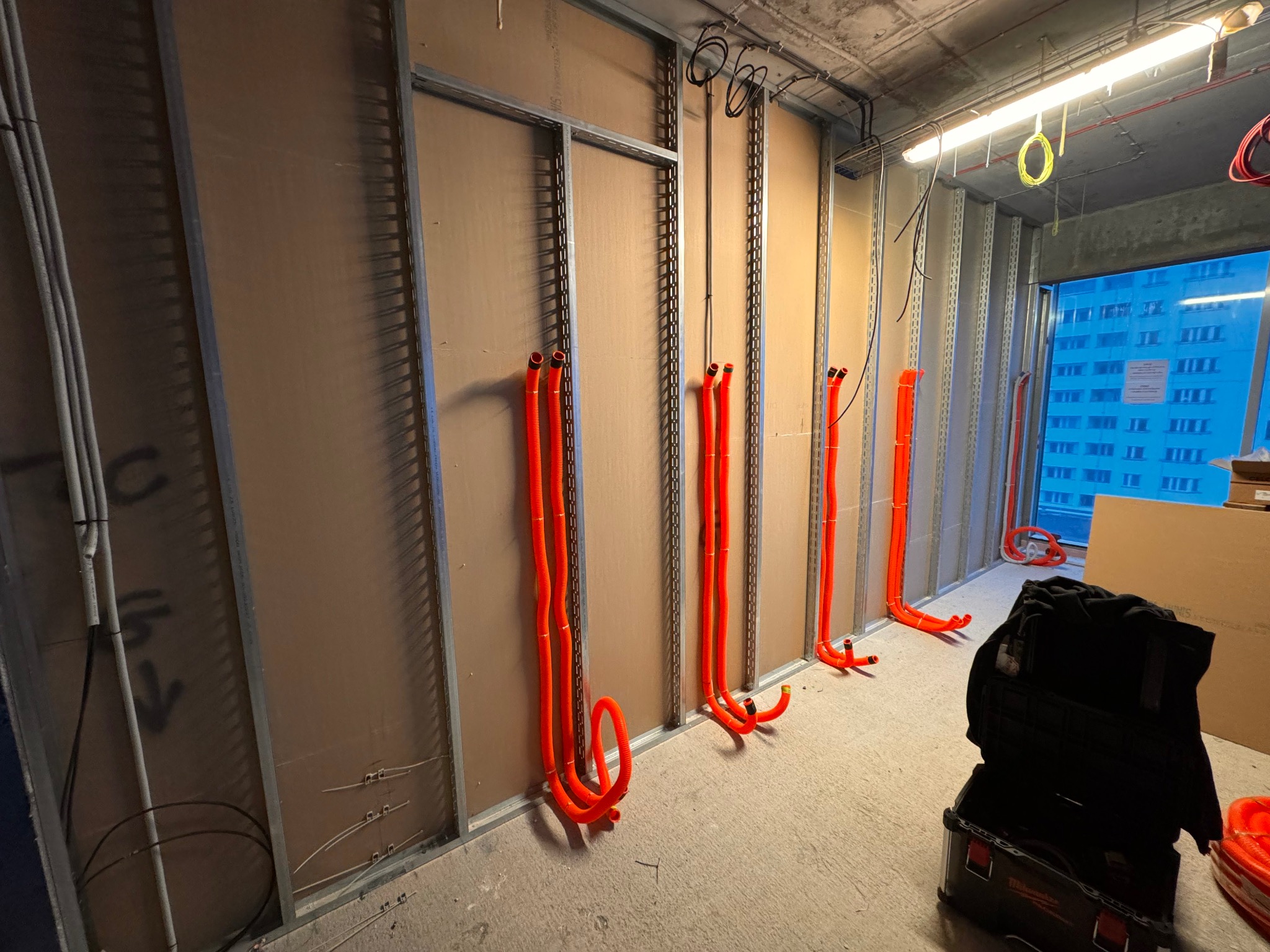 Montaż pomarańczowych rur instalacyjnych wewnątrz metalowej konstrukcji ścian działowych, widoczna skrzynka narzędziowa w tle.