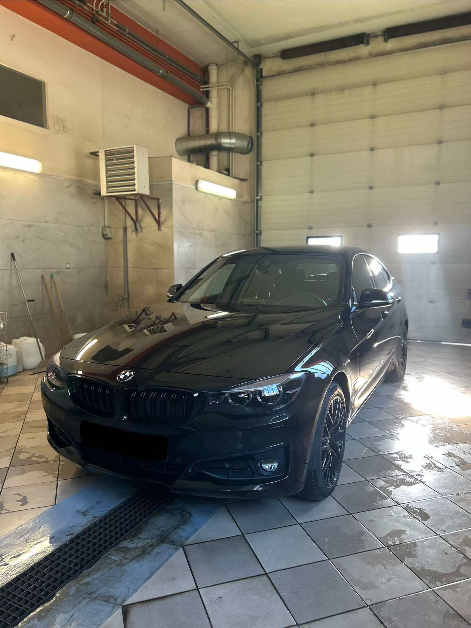 Czarny samochód BMW w myjni, widoczna mokra podłoga i kratka odpływowa, oświetlenie sufitowe.