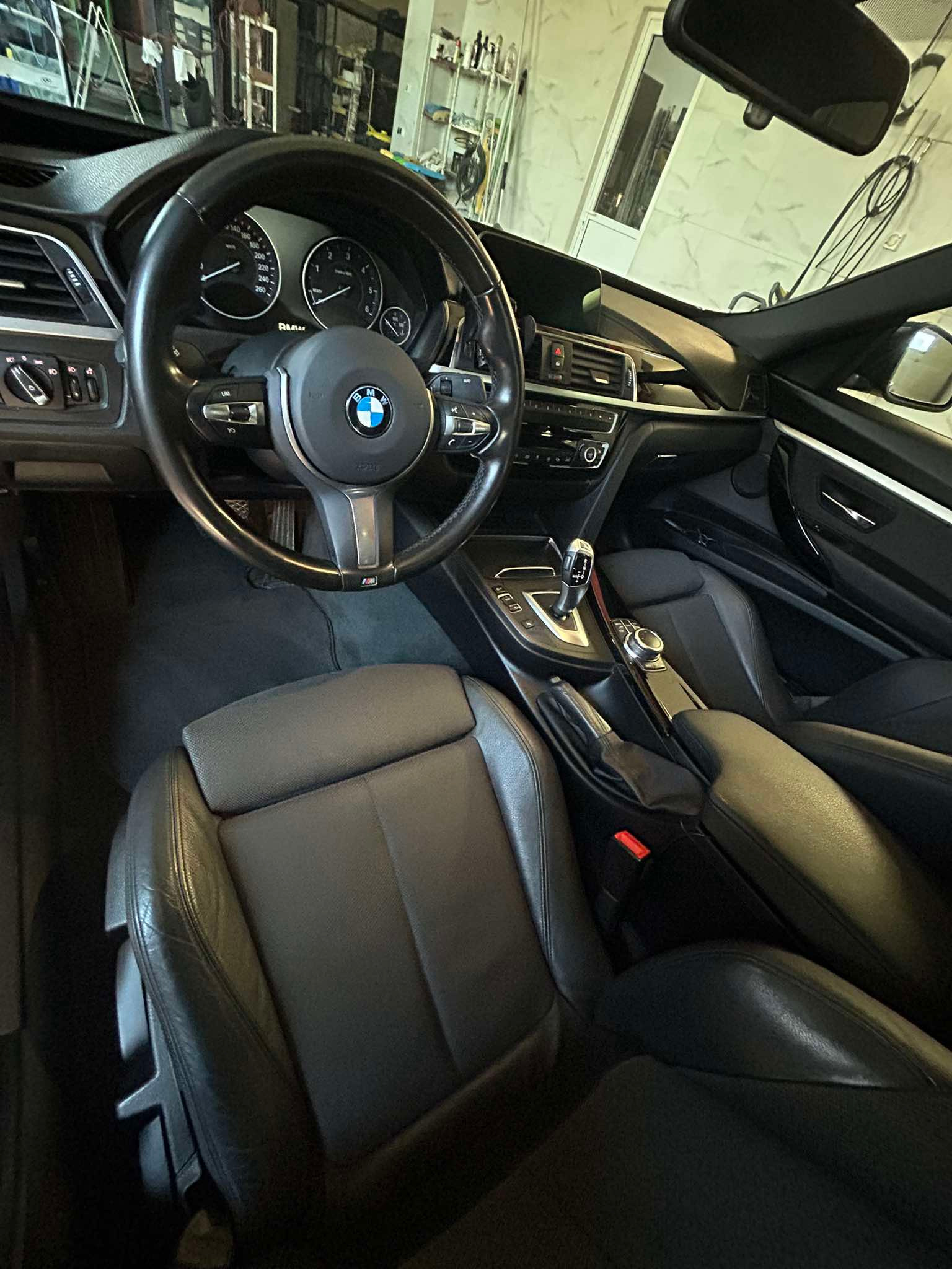 Wnętrze samochodu BMW, widok z przodu na deskę rozdzielczą, kierownicę z logo, skórzane fotele i konsolę środkową. Widoczny fragment szyby przedniej i lusterka wstecznego.