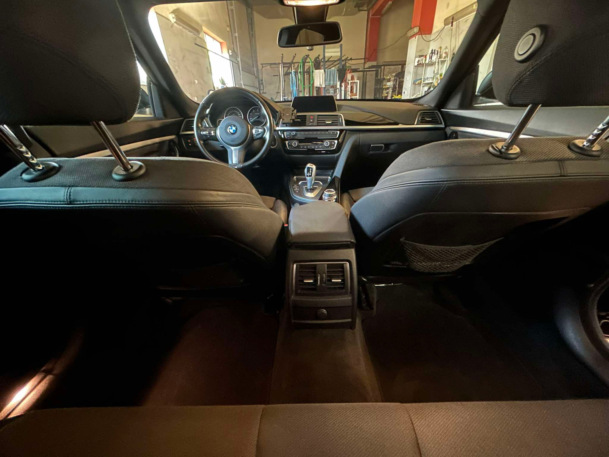Wnętrze samochodu BMW widziane z tylnych siedzeń, z widoczną deską rozdzielczą, kierownicą z logo marki i dźwignią zmiany biegów, wnętrze czyste, w tle widoczna myjnia samochodowa.