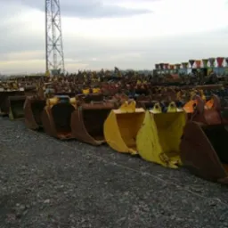 Rząd używanych łyżek do koparek w różnych kolorach (żółty, brązowy) na żwirowym terenie. W tle widoczna wieża telekomunikacyjna i inne maszyny budowlane.