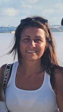 Uśmiechnięta kobieta w białym podkoszulku i okularach na głowie, stojąca na tle jasnego nieba i wody. Widoczny pasek z napisem 'Mickey'.