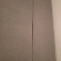 Fragment szarej, tekstylnej tapicerki kanapy, widoczny szew i narożnik poduszki oparcia.