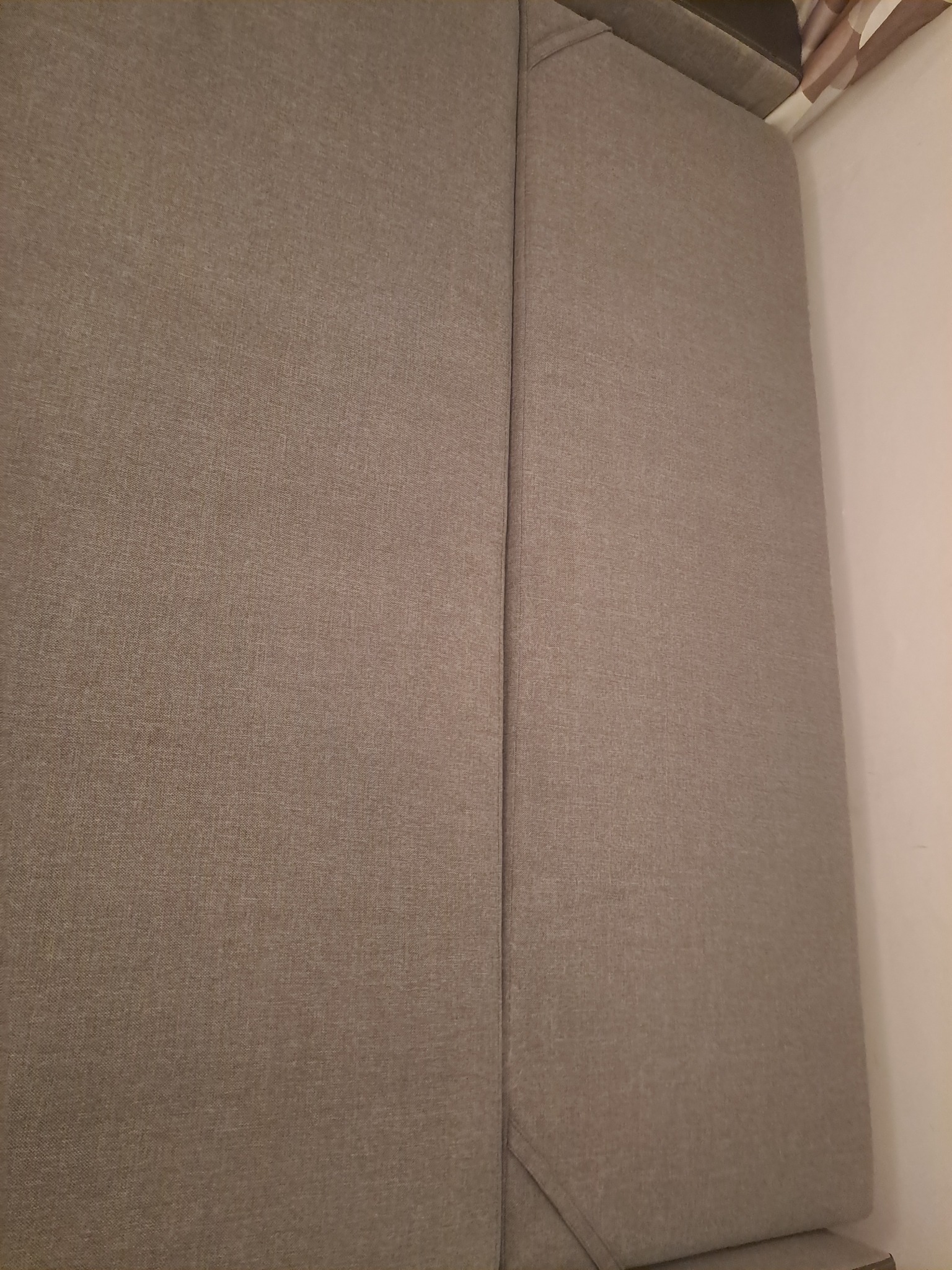 Fragment szarej, tekstylnej tapicerki kanapy, widoczny szew i narożnik poduszki oparcia.