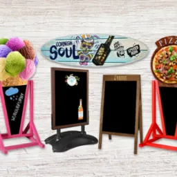 Różnorodne tablice reklamowe: różowa z lodami i napisem 'Wodoodporny', czarna z logo firmy i napojem, drewniana z logo Dunkin' Donuts, czerwona z pizzą, oraz deska surfingowa z napisem 'Cornish...