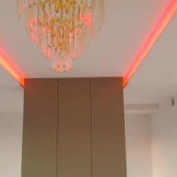 VASYL HNATUSHKO VIVERE INMOMENTO - Kryształowy żyrandol z złoconym wykończeniem, oświetlony czerwonymi diodami LED w suficie, nad zabudową meblową w kolorze taupe.