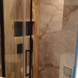 VASYL HNATUSHKO VIVERE INMOMENTO - Nowoczesna kabina prysznicowa z czarnymi elementami i drewnopodobnymi płytkami na ścianach, z kwadratową deszczownicą w ochronnej folii i odpływem liniowym w podłodze.