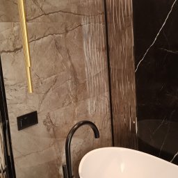 VASYL HNATUSHKO VIVERE INMOMENTO - Elegancka łazienka z wolnostojącą białą wanną, czarną armaturą i okładziną ścienną z marmuru w odcieniach brązu i czerni, oświetlona złotą lampą wiszącą.