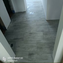 VASYL HNATUSHKO VIVERE INMOMENTO - Ułożone szare płytki podłogowe imitujące drewno w przejściu, widoczne ślady po czyszczeniu, surowe ściany w trakcie prac wykończeniowych.