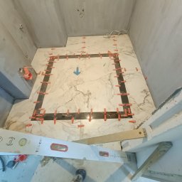 VASYL HNATUSHKO VIVERE INMOMENTO - Montaż marmurowych płytek podłogowych w łazience, widoczne krzyżyki dystansowe i poziomica, surowe ściany w tle.