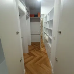 Wnętrze garderoby z białymi szafami i parkietem ułożonym w jodełkę, widok z perspektywy wejścia.