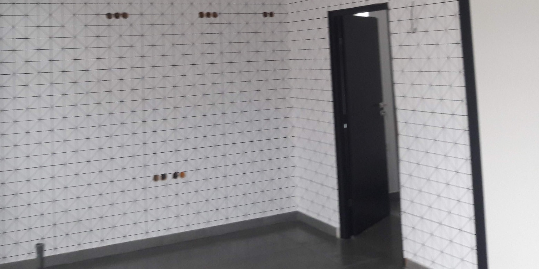 Ściana wyłożona białymi płytkami z czarnymi liniami fugi, widoczne otwory na gniazdka elektryczne i czarne drzwi w tle.