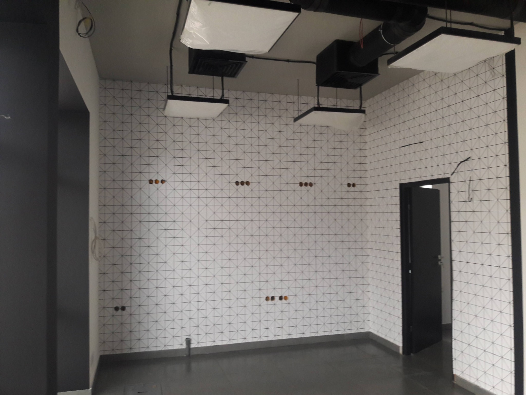 Wnętrze w trakcie prac wykończeniowych: ściany wyłożone białą mozaiką z czarnym wzorem geometrycznym, widoczne otwory na gniazdka elektryczne, czarne ramy drzwi i sufit z zamontowanym oświetleniem.