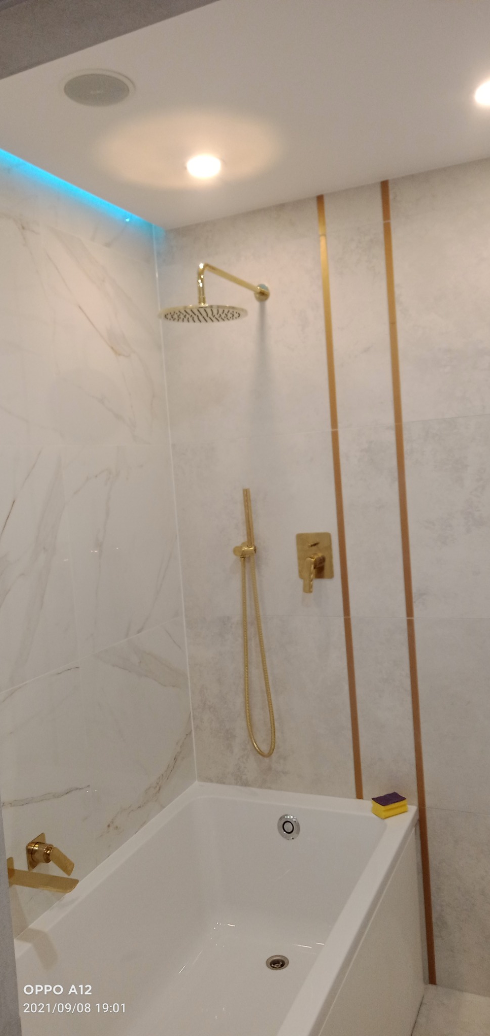 Nowoczesna łazienka z białą wanną, marmurowymi płytkami i złotą armaturą, akcentowana niebieskim oświetleniem LED podsufitowym. Na wannie leży żółto-fioletowa gąbka do czyszczenia.