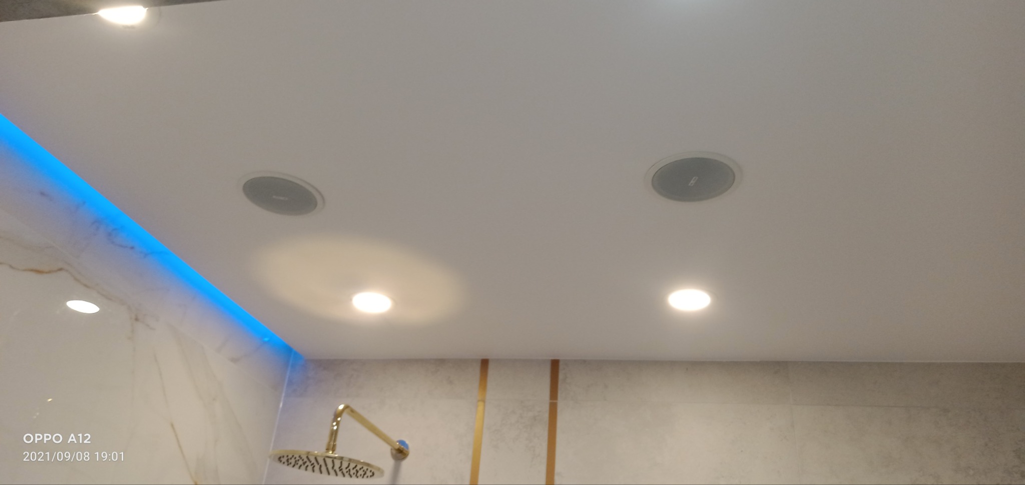 Nowoczesna łazienka z podświetleniem LED na suficie i marmurowymi płytkami ściennymi, widoczne głośniki sufitowe i złota deszczownica.