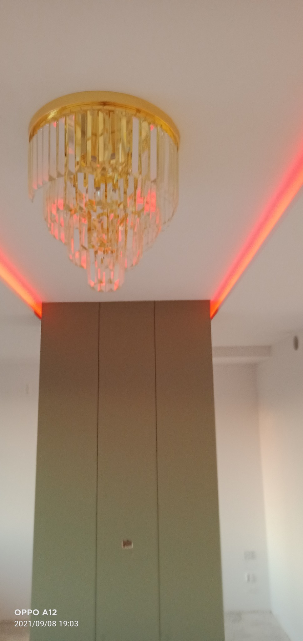 Kryształowy żyrandol z złoconym wykończeniem, oświetlony czerwonymi diodami LED w suficie, nad zabudową meblową w kolorze taupe.