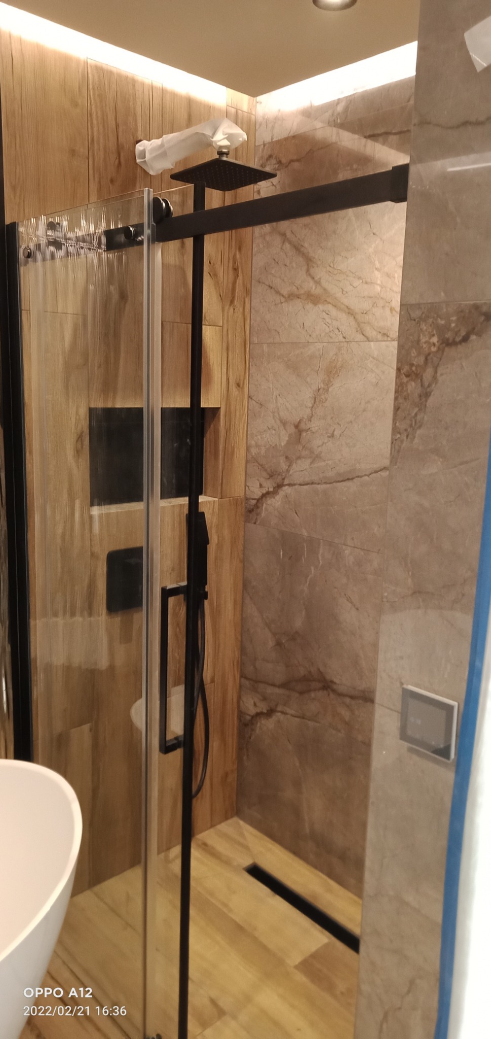 Nowoczesna kabina prysznicowa z czarnymi elementami i drewnopodobnymi płytkami na ścianach, z kwadratową deszczownicą w ochronnej folii i odpływem liniowym w podłodze.