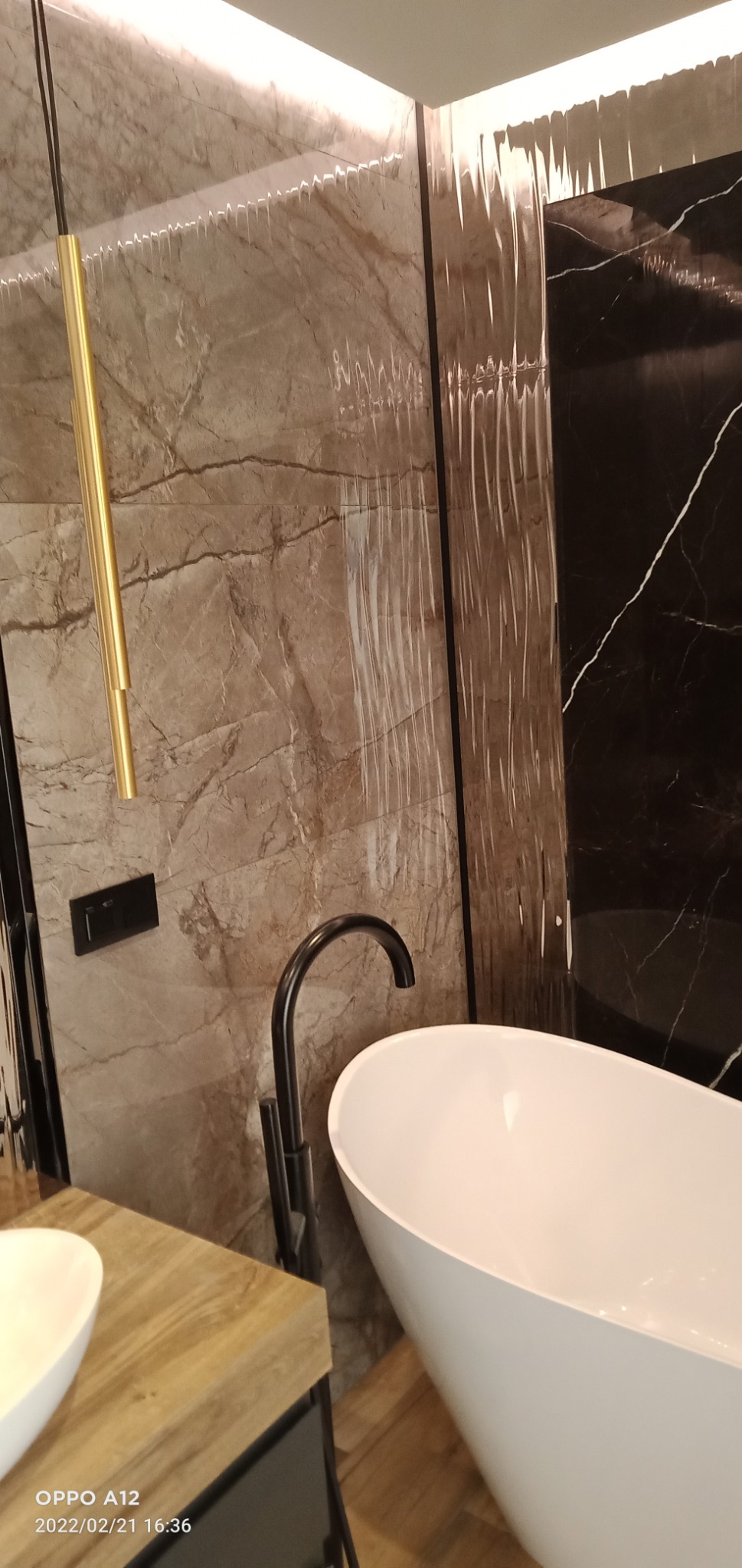 Elegancka łazienka z wolnostojącą białą wanną, czarną armaturą i okładziną ścienną z marmuru w odcieniach brązu i czerni, oświetlona złotą lampą wiszącą.