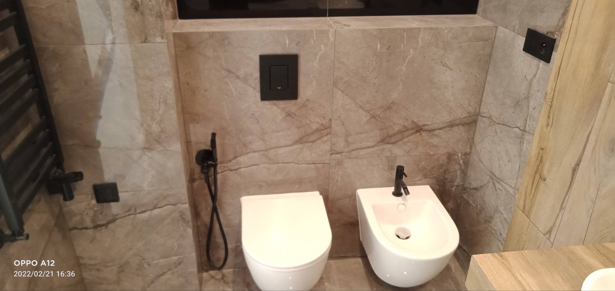 Nowoczesna łazienka z marmurowymi płytkami, wiszącą toaletą i bidetem z czarną armaturą, oraz grzejnikiem drabinkowym w kolorze czarnym