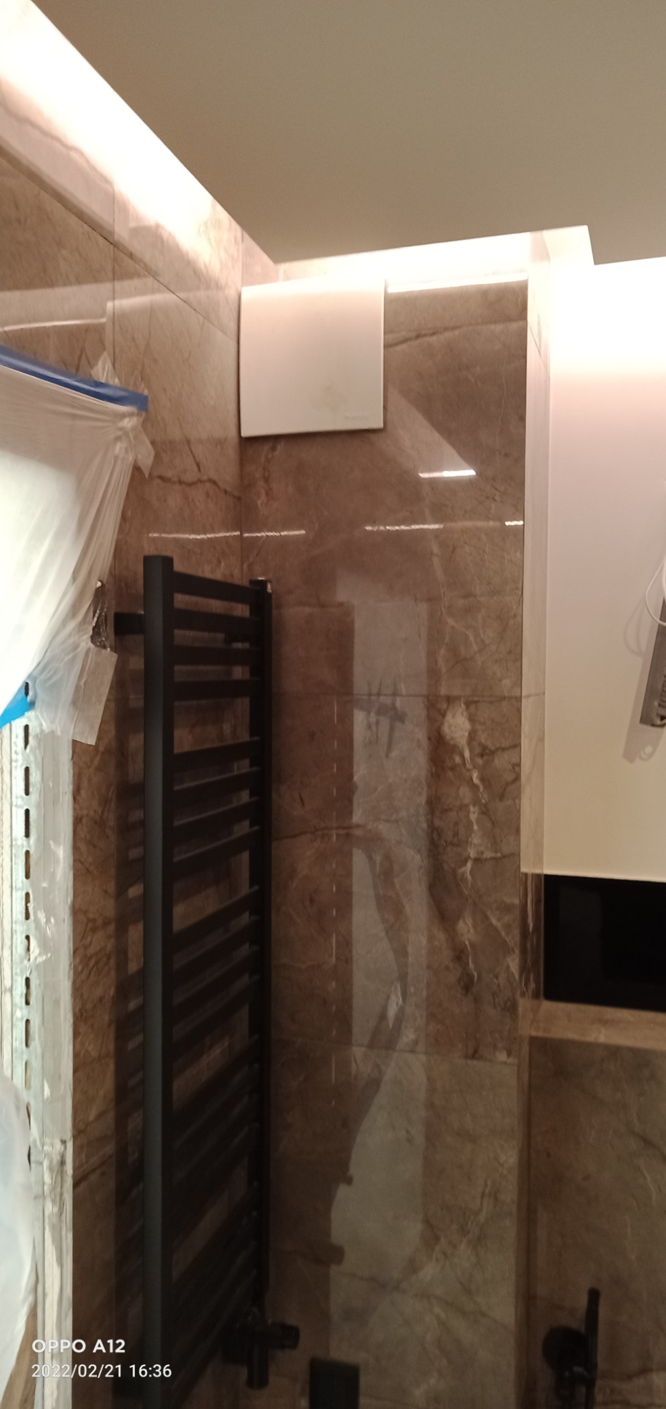 Częściowo zakryty folią grzejnik łazienkowy w kolorze czarnym, zamontowany na ścianie z płytek imitujących marmur, widoczny fragment wentylatora sufitowego.