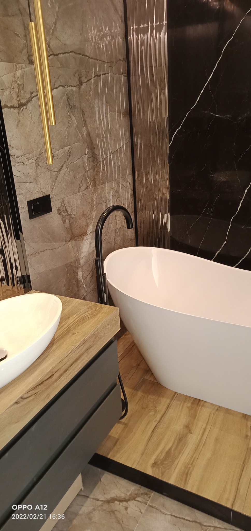 Nowoczesna łazienka z wolnostojącą białą wanną, czarną armaturą, umywalką nablatową na drewnopodobnej szafce i marmurowymi płytkami na ścianach w odcieniach szarości i czerni.