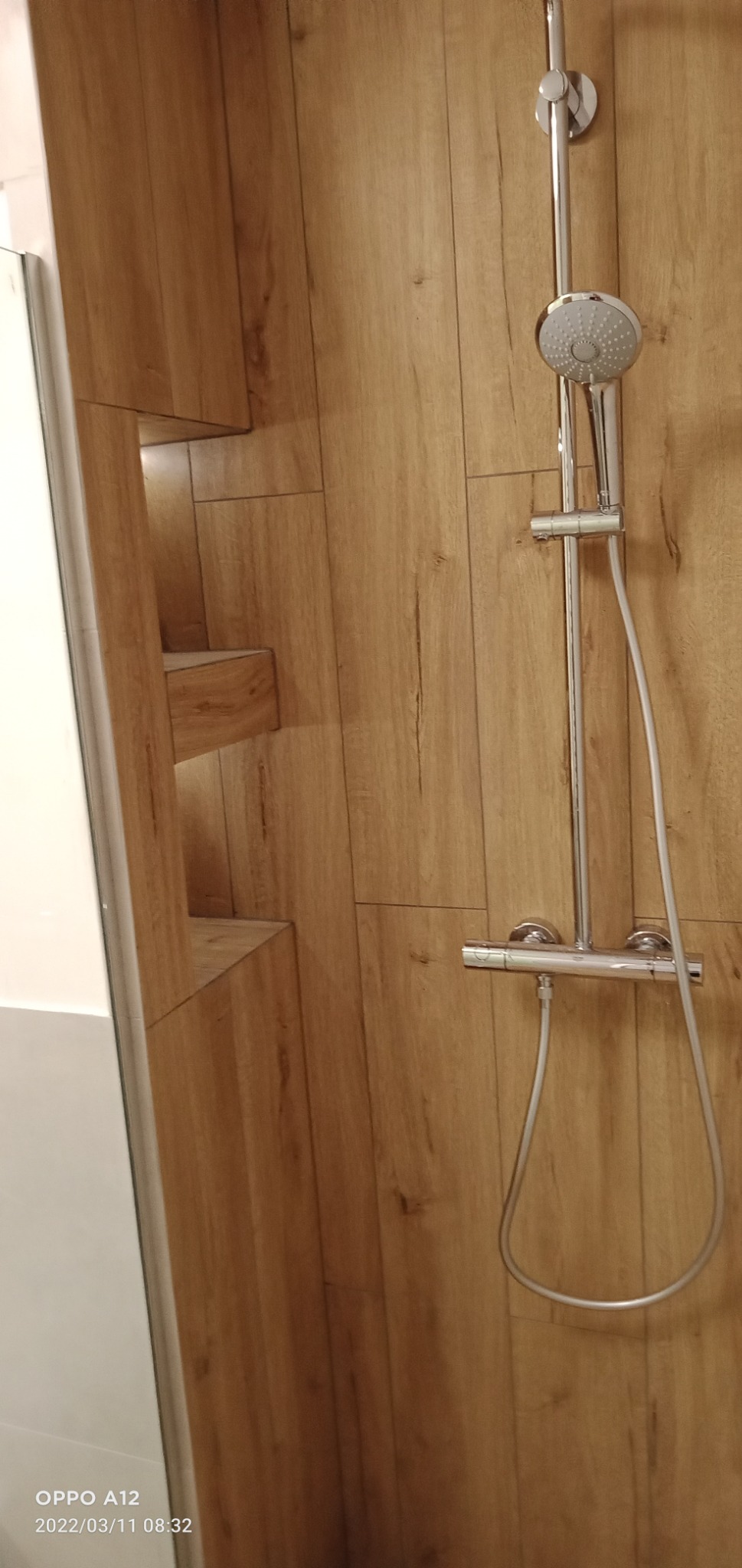 Kabina prysznicowa z drewnopodobnymi płytkami na ścianach, chromowana bateria i deszczownica, półki wnękowe wykończone w tym samym stylu.