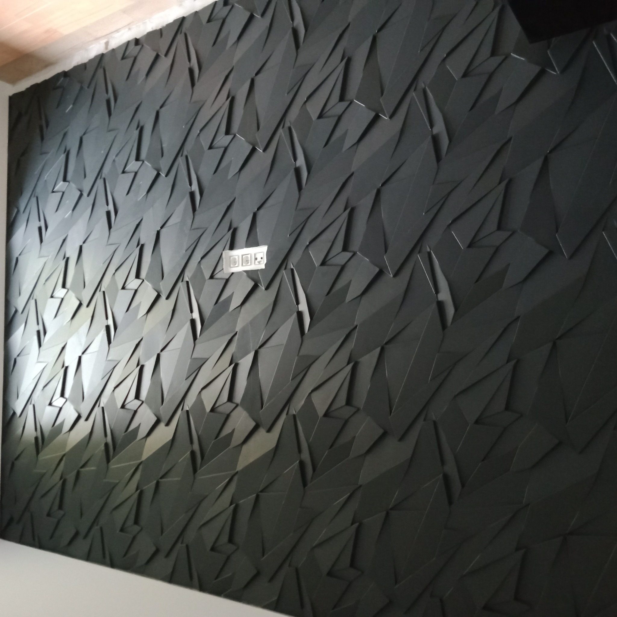 Czarna ściana z trójwymiarowymi panelami dekoracyjnymi o geometrycznym wzorze, z widocznym gniazdkiem elektrycznym po lewej stronie