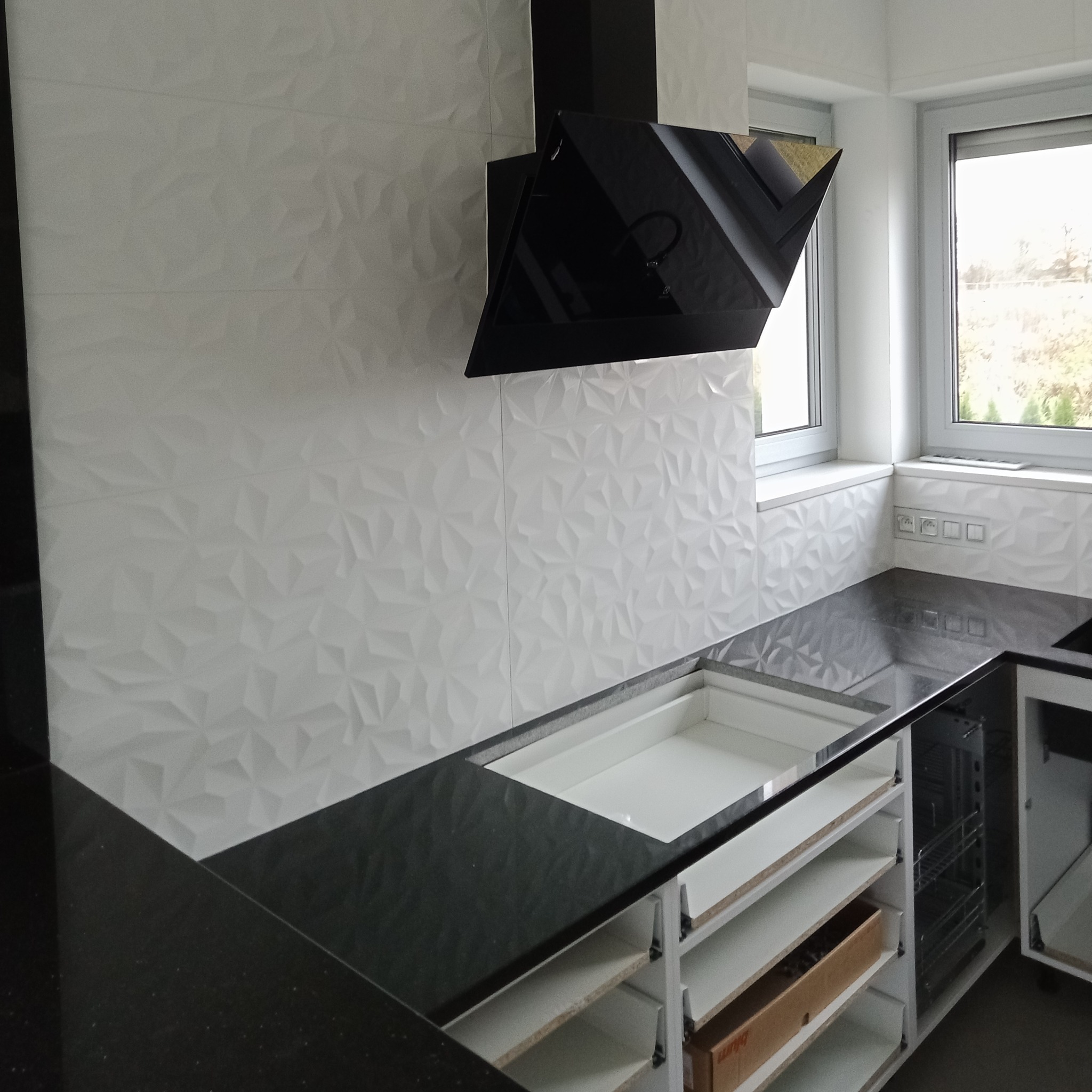 Nowoczesna kuchnia w trakcie montażu: czarny blat, biała, trójwymiarowa ściana z płytek, czarny okap i okno z widokiem na zieleń.