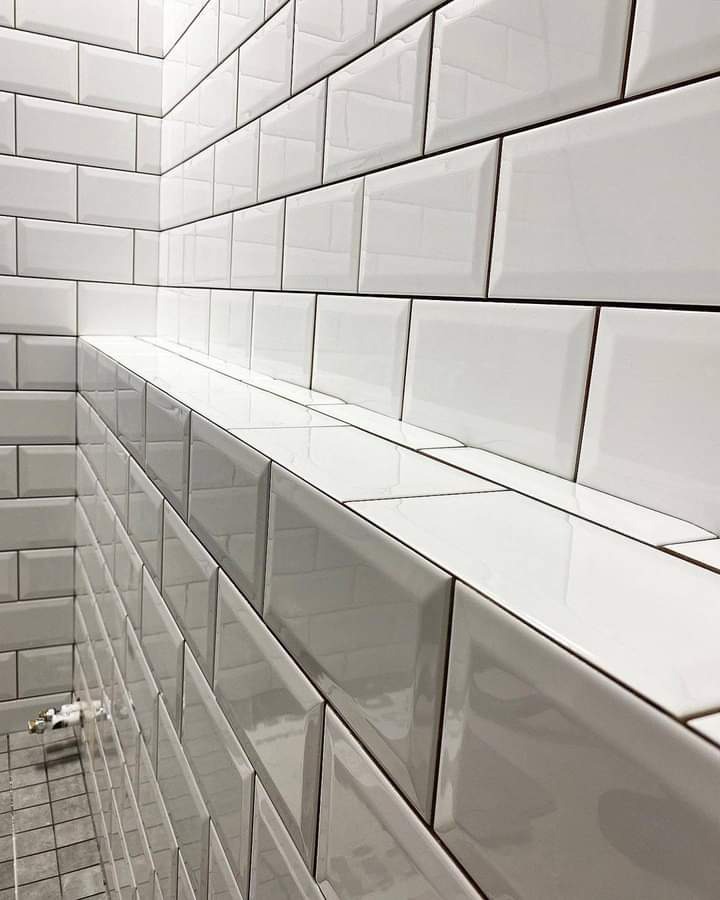 Białe, błyszczące płytki typu metro ułożone na ścianie i półce w łazience, widoczna szara fuga i fragment podłogi z odpływem liniowym.