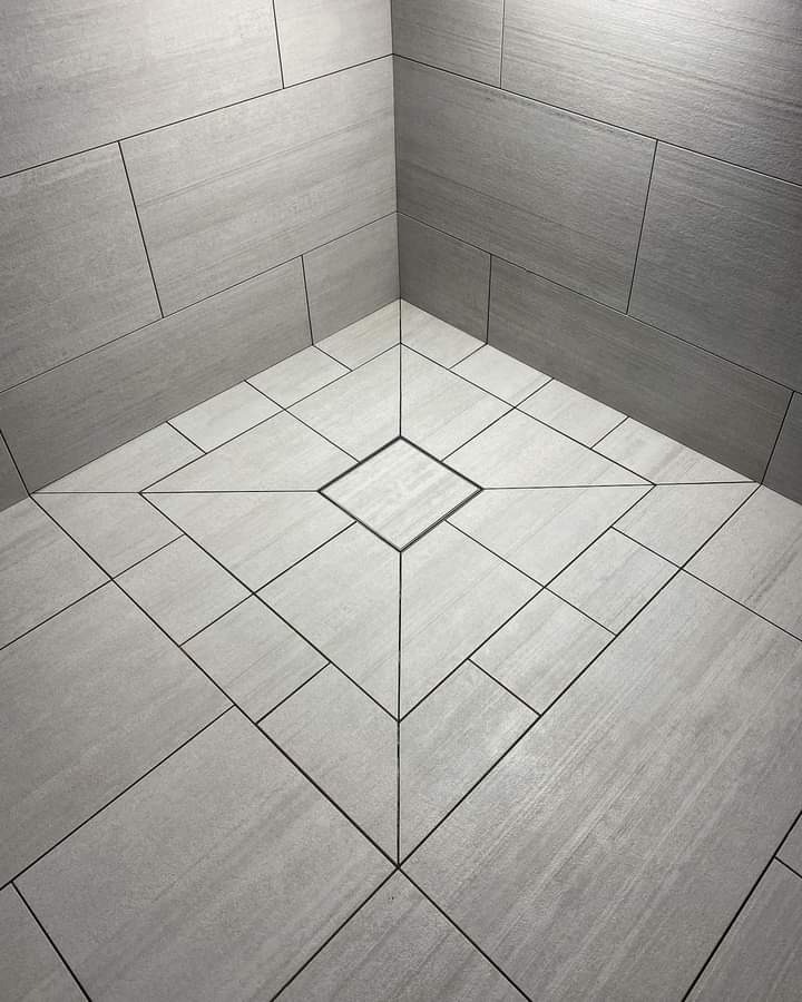 Wykończony narożnik łazienki z szarymi płytkami ułożonymi w geometryczny wzór, z centralnym odpływem liniowym w kształcie kwadratu.