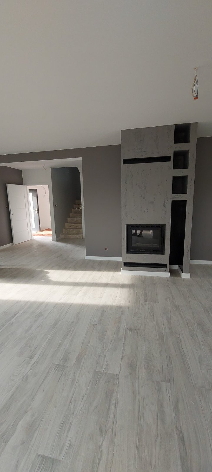 Pusty pokój z szarą podłogą imitującą drewno, szarymi ścianami i nowoczesnym kominkiem z półkami. W tle widoczne schody i otwarte drzwi.