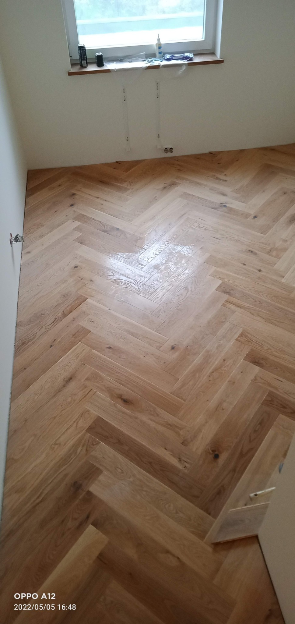 Parkiet ułożony we wzór jodełki francuskiej, częściowo pokryty lakierem, widoczne niedoskonałości przy ścianie, białe ściany i okno w tle.