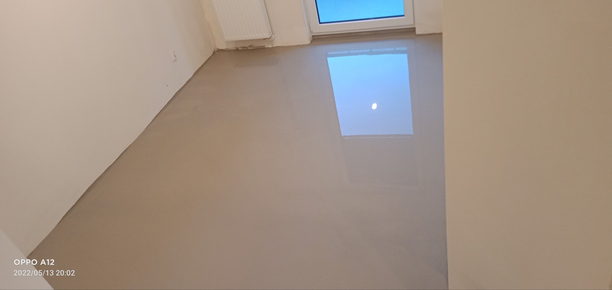 Wylany, mokry samopoziomujący podkład podłogowy w pokoju z białymi ścianami i grzejnikiem. W podłodze odbija się okno dachowe.