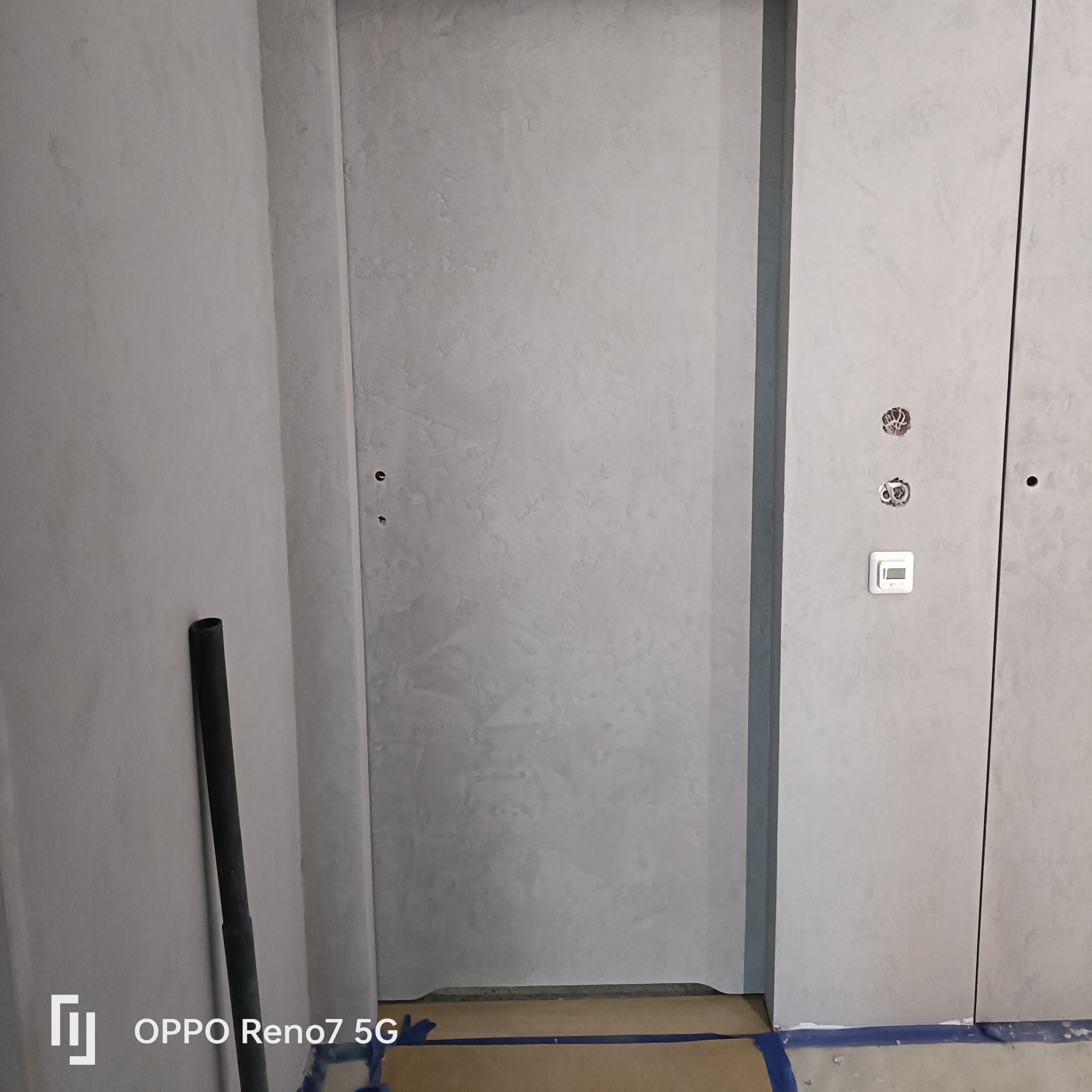 Świeżo pomalowana szpachlowana ściana w odcieniach szarości z widocznymi otworami na gniazdka elektryczne i termostat, zabezpieczona taśmą malarską przy podłodze.