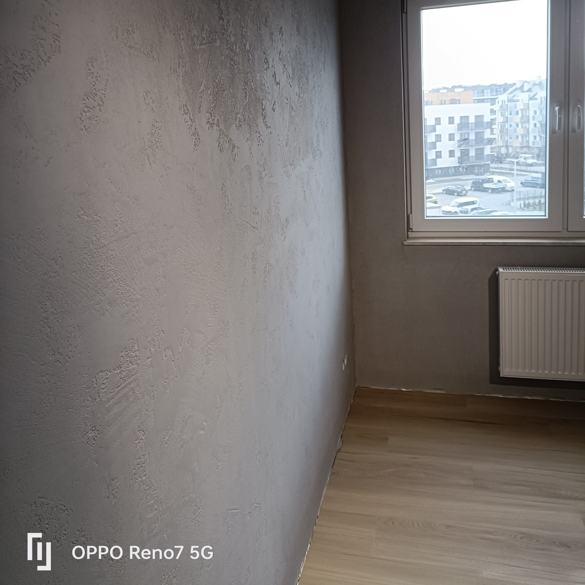 Ściana z dekoracyjną szarą strukturą, widok na okno z osiedlem mieszkaniowym, grzejnik i fragment podłogi z paneli.