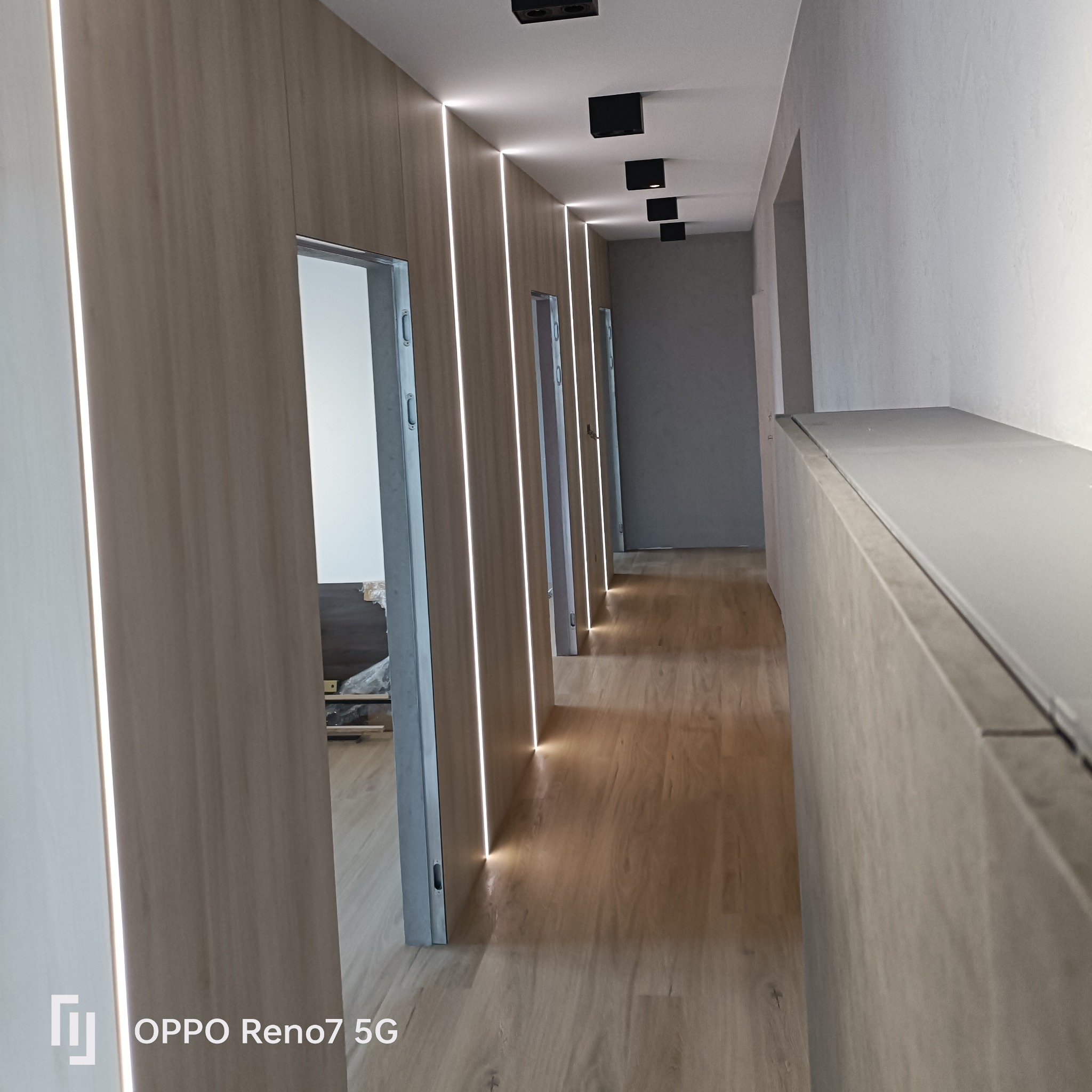 Długi korytarz z drewnianą podłogą i ścianami z wbudowanym oświetleniem LED, widoczne drzwi bez futryn i czarne lampy sufitowe.